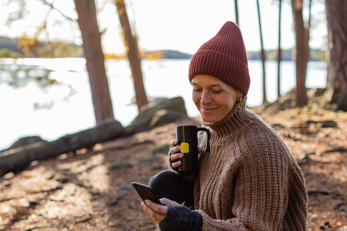 Dame drikker te i skogen og ser på mobilen