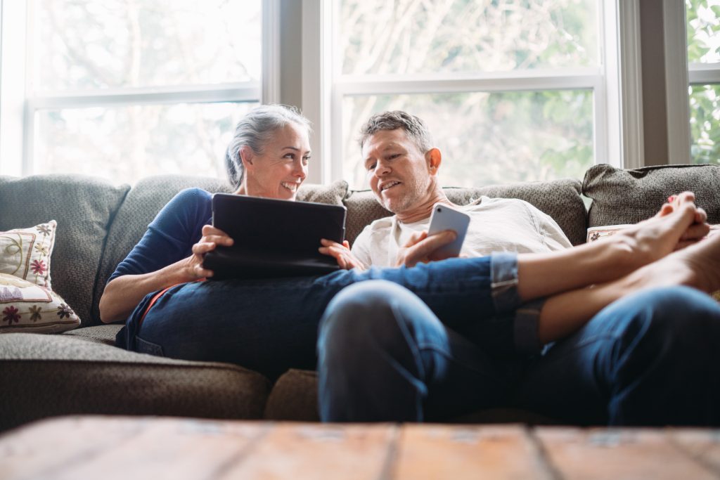 Mann og kone ser på nettbrett og mobil i sofaen